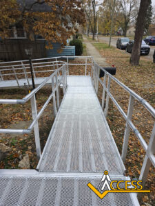 A metal wheelchair ramp with handrails leads down to a sidewalk lined with autumn leaves. Houses and trees can be seen along the street. The Access company logo is in the lower right corner.