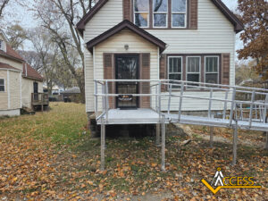 A beige house with brown trim has a metal wheelchair ramp leading to the front door. Fallen autumn leaves cover the yard. The Access company logo is in the lower right corner.