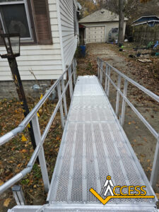 A metal wheelchair ramp with railings leads up to the side entrance of a house. Leaves are scattered on the ground, and the word ACCESS with a yellow logo appears in the bottom right corner.