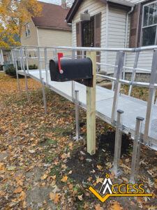 A silver metal wheelchair ramp leads to a house, passing next to a black mailbox on a wooden post. Fallen autumn leaves cover the ground, and the Access logo is in the bottom right corner.
