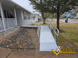 An aluminum wheelchair ramp leads from a sidewalk to the front porch of a house. Two people stand on the porch. The yard has autumn leaves and a sloped rock bed. The Access logo is in the lower right corner.