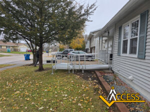 A metal wheelchair ramp leads from the sidewalk to the front door of a house, passing beside a tree. Several cars are parked nearby, and the Access company logo is in the lower right corner.