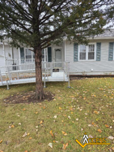 A white house with blue shutters has a metal wheelchair ramp leading to the front door. A large tree stands in the lawn, and the ground is scattered with fallen leaves. The ACCESS logo is visible in the corner.