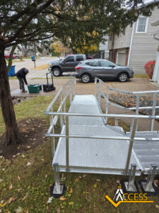 A newly installed aluminum wheelchair ramp leads from a sidewalk to a house. A person works near a toolbox in the yard. Cars are parked on the street, and autumn leaves are scattered on the ground. Access company logo in bottom right.