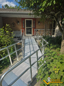 A metal wheelchair ramp with railings leads to the front door of a house with a red trim. A garden surrounds the ramp, and a bench sits on the porch. ACCESS logo appears in the corner.