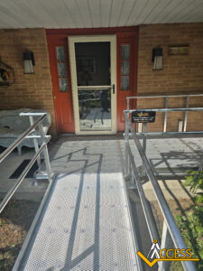 A metal wheelchair ramp leads to a red front door with glass panels on a brick house. A white bench sits to the left, and the word “Access” with a logo is visible in the lower right corner.