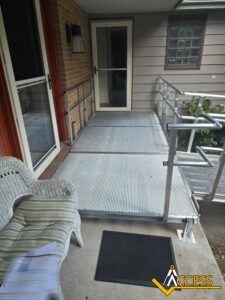 A metal wheelchair ramp with railings leads to a front door, providing accessible entry to a house. A wicker chair with a cushion and a black mat sit nearby. The Access logo is visible in the bottom right corner.