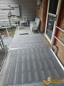 A metal wheelchair ramp leads to a porch with wicker chairs and a small table. The house has brown brick walls and utility boxes are mounted nearby. The Access logo is in the corner of the image.