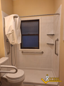 Accessible bathroom with a walk-in shower, white tile walls, grab bars, a shower curtain partially pulled back, a toilet with a support rail, and built-in corner shelves. The ACCESS logo is in the bottom right corner.