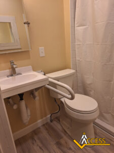 A small accessible bathroom with a wall-mounted sink, a toilet with a white support rail attached, a mirror, a shower with a white curtain, light-colored walls, and wood-patterned flooring.
