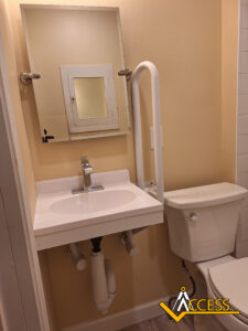 Small accessible bathroom with a wall-mounted sink, exposed plumbing beneath, a mirror above the sink, a toilet next to the sink, and a white grab bar between the toilet and sink. ACCESS logo is in the lower right corner.