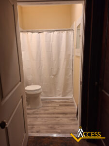Small bathroom with light-colored walls and flooring, featuring a white toilet and a shower with a white curtain. The photo is taken from the doorway. The ACCESS logo is visible in the bottom right corner.