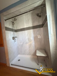 A walk-in shower with gray and white tiled walls, two showerheads, a handheld shower, a grab bar, and a white shower chair. ACCESS logo is in the bottom right corner.
