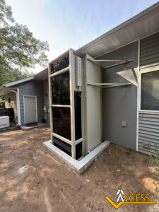 An outdoor residential wheelchair lift with glass panels is installed next to a house with gray siding. The lift sits on a concrete base, and the ACCESS logo is visible in the bottom right corner. Trees are in the background.