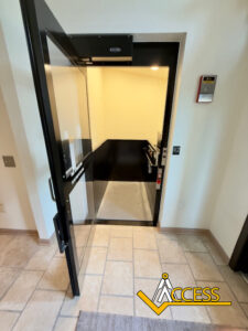 A small elevator with a black frame and open glass door is visible. The elevators interior is brightly lit, and the ACCESS logo appears in the bottom right corner of the image.
