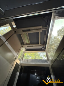 View from inside an elevator, looking up at the open ceiling panel with visible mechanical parts and natural light coming through glass panels; trees are visible outside. The Access logo is in the bottom right corner.
