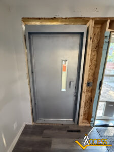 A gray metal door with a narrow vertical window is installed in an unfinished doorway, surrounded by exposed wood framing. The Access logo appears in the bottom right corner.
