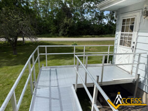 A metal wheelchair-accessible ramp with railings leads to the back door of a light blue house, surrounded by green grass and trees. The ACCESS logo is visible in the bottom right corner.