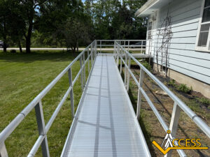 A metal wheelchair ramp leads up to the side entrance of a light blue house. The ramp is surrounded by grass and trees, with the ACCESS company logo in the bottom right corner.