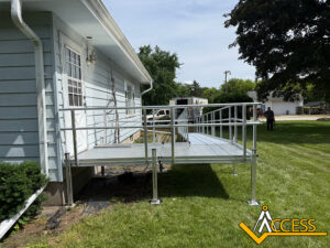 A metal accessibility ramp with railings is attached to the side entrance of a light blue house, providing access from the doorway to the grassy yard. The Access company logo is in the bottom right corner.