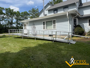 A long aluminum wheelchair ramp leads to the side entrance of a light blue house, surrounded by grass and trees. An Access logo is in the bottom right corner.