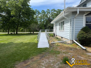 A metal wheelchair ramp leads up to the side entrance of a light blue house with white trim, surrounded by grass, trees, and bushes. The Access company logo is in the bottom right corner.