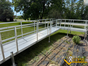 A metal wheelchair ramp with handrails extends over grass in a yard, leading to a garden area. Trees and open green space are visible in the background. The Access logo is in the lower right corner.