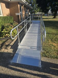 A metal wheelchair ramp with railings leads from a sidewalk up to the entrance of a brick house, passing by landscaped bushes and a grassy yard.