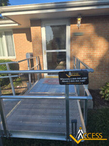 A metal wheelchair ramp leads to a glass door entrance of a brick building. A sign displays contact information for ACCESS, a ramp provider, with their logo in the lower right corner.