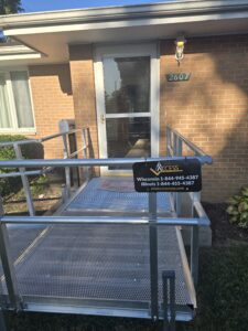 A metal wheelchair ramp leads to the front door of a brick building with the address number 2607. A sign on the ramp displays contact information for ACCESS services in Wisconsin and Illinois.