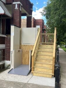 A wooden staircase with a handrail is next to an outdoor wheelchair lift installed on a concrete sidewalk, providing accessible entry to a brick building. Trees and a street are visible in the background.
