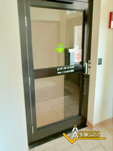 A glass door with a black frame, a push bar, and caution stickers stands in a hallway. A tiled floor and beige walls are visible. The word Access with a yellow logo appears in the bottom right corner.