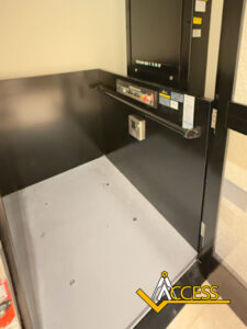 A black platform wheelchair lift with control buttons and safety railings on the sides, installed indoors next to a wall. The logo Access is visible in the bottom right corner.