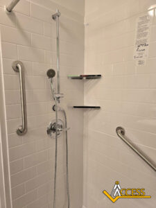 A white tiled shower with a handheld showerhead, two stainless steel grab bars, two corner shelves, and a visible ACCESS logo in the bottom right corner.