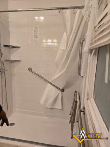 Accessible shower with white tiles, a handheld showerhead, a grab bar, a fold-down shower chair, corner shelves, and a partially open white shower curtain. The ACCESS logo is visible in the lower right corner.