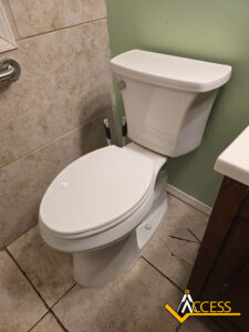 White toilet with closed lid in a bathroom with beige tiled walls and floor, pale green wall behind, and a wooden cabinet to the right. ACCESS logo is visible in the bottom right corner.