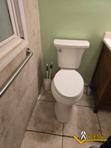 A bathroom with a white toilet, a grab bar on a tiled wall, a toilet brush set in the corner, and a sink cabinet. The floor is tiled and a purple spray bottle is near the cabinet.