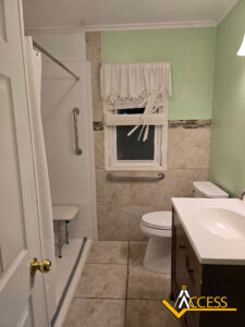 Accessible bathroom with a walk-in shower featuring a grab bar and shower seat, a window with a white curtain, a toilet, and a vanity with a sink. ACCESS logo is in the bottom right corner.