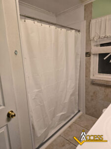 A bathroom with a white shower curtain on a rod, beige tiled walls, a white door with a gold handle, a window with a curtain, and the Access logo in the bottom right corner.