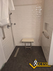 Accessible shower with white tiled walls, a foldable shower seat, handheld showerhead, and grab bars on both sides. The floor is dark, and there is a logo with the word ACCESS in the bottom right corner.