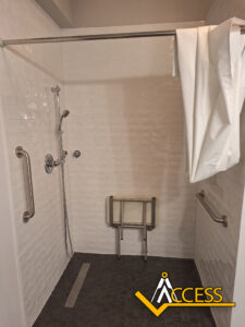 An accessible shower with a wall-mounted folding seat, grab bars on two sides, a handheld showerhead, a white curtain, and the ACCESS logo in the bottom right corner.
