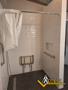 Accessible shower with white subway tile walls, a grab bar, a foldable shower seat, a shower curtain, and a recessed shelf. The Access logo is visible in the bottom right corner.