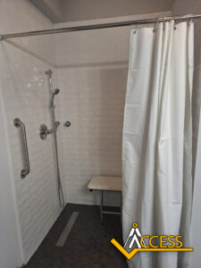 Accessible walk-in shower with white tile walls, a handheld shower head, horizontal grab bars, a shower bench, dark hexagonal floor tiles, and a white shower curtain. Access logo appears in the bottom right corner.