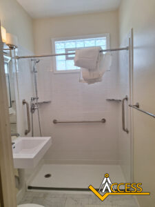 Accessible shower with grab bars, fold-down shower seat, handheld showerhead, wall-mounted sink, towels hanging on the curtain rod, and a frosted glass window. ACCESS logo is in the bottom right corner.