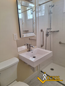 Accessible bathroom with a wall-mounted sink, angled mirror, grab bars, handheld showerhead, and a walk-in shower; ACCESS logo is in the corner.