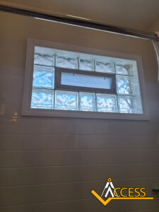 A glass block bathroom window with a small vent in the center, surrounded by white tiled walls. The Access logo appears in the lower right corner.
