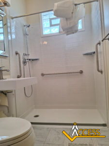 A spacious, accessible walk-in shower with grab bars, a handheld showerhead, a fold-down shower seat, and a towel draped over the shower rod. The Access logo is visible in the bottom right corner.
