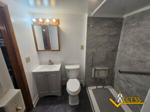 A bathroom with gray tile flooring, a vanity with a sink and mirror, a toilet, and an accessible shower with grab bars and a fold-down seat. The Access company logo is in the bottom right corner.