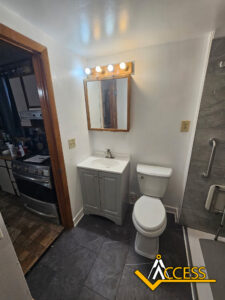 Small bathroom with a mirror above a gray vanity, sink, and toilet. The floor is dark tile, and part of a stove from the adjoining kitchen is visible. Access logo in the lower right corner.
