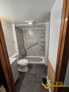 A modern accessible bathroom with gray tile, a walk-in shower with grab bars, a handheld showerhead, corner shelves, a toilet, and a small white sink. The Access company logo is in the bottom right corner.